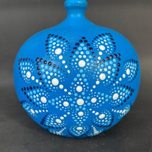 Puede incluir: Un jarrón redondo de cerámica azul pintado a mano con un cuello pequeño. El jarrón está decorado con un patrón de puntos blancos y azul oscuro. Los puntos están dispuestos en un diseño simétrico en la superficie del jarrón.