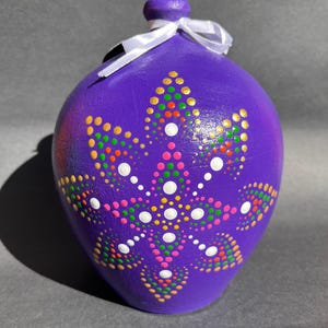 Puede incluir: Una hucha de cerámica morada con una cinta blanca atada al cuello. La hucha está decorada con un colorido patrón de puntos en rosa, blanco, amarillo, verde y naranja. La hucha tiene forma redondeada.