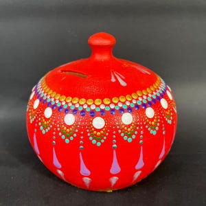 Puede incluir: Una hucha roja de cerámica con tapa y ranura para monedas. La hucha está decorada con un patrón de puntos blancos, dorados, turquesas y morados. El diseño es simétrico y cubre toda la superficie.