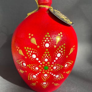 Puede incluir: Adorno de cerámica roja pintado a mano con una cinta dorada. El adorno presenta un diseño floral con patrones de puntos blancos, dorados y verdes. El adorno tiene forma redonda y una pequeña etiqueta.