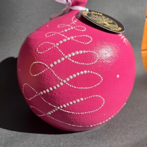 Puede incluir: Un adorno esférico rosa brillante con remolinos blancos punteados y una cinta blanca. Se adjunta una etiqueta negra con texto dorado. El adorno es probablemente para la decoración navideña.