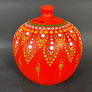 Puede incluir: Una hucha redonda de cerámica roja con una ranura en la parte superior. La hucha está decorada con un patrón de puntos blancos, amarillos y dorados. Tiene un pequeño pomo redondo en la parte superior.