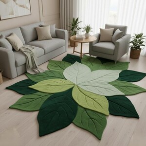 May include: A leaf-shaped rug with a central flower design. The rug features a variety of green shades, from dark to light, creating a naturalistic look. The rug is placed in a living room setting with gray furniture and plants.