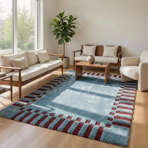 May include: A rectangular area rug featuring a light blue central field surrounded by a border of alternating light blue and deep red rectangular blocks. The rug has a thick, textured pile and is positioned on a light-colored wooden floor.