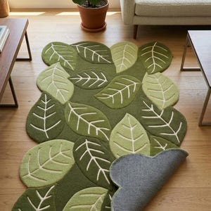 May include: A rectangular area rug featuring a pattern of overlapping green leaves in various shades of olive and lime. The rug has a tufted texture with white stitched leaf veins. One corner of the rug is folded over to reveal a gray backing.