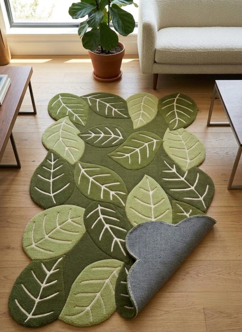 May include: A rectangular area rug featuring a pattern of overlapping green leaves in various shades of olive and lime. The rug has a tufted texture with white stitched leaf veins. One corner of the rug is folded over to reveal a gray backing.