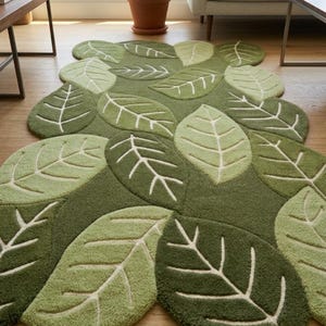 May include: A tufted area rug featuring an organic, irregular shape composed of overlapping leaf motifs in various shades of forest, olive, and sage green. Each leaf includes a central vein and branching lines in a contrasting cream color.