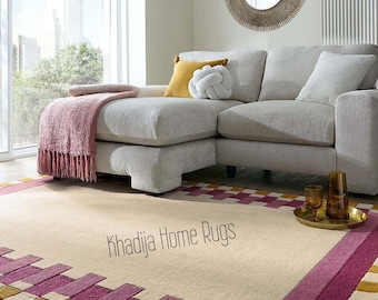 Op de afbeelding: Beige vloerkleed met een geometrisch patroon in roze en goud. Het vloerkleed ligt in een woonkamer met een grijze hoekbank, een roze deken en een decoratieve spiegel. De tekst "Khadija Home Rugs" is op het vloerkleed gedrukt.