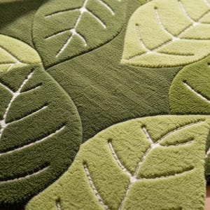 May include: A tufted rug featuring a repeating pattern of overlapping leaves in various shades of green, including olive, lime, and forest green. Each leaf shape is defined by a recessed white stitched vein detail, creating a textured surface.