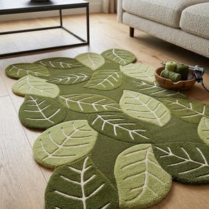 May include: A tufted area rug shaped like a cluster of overlapping leaves in various shades of olive, forest, and sage green. The rug features a raised, textured surface with cream-colored stitched vein details on each leaf segment.