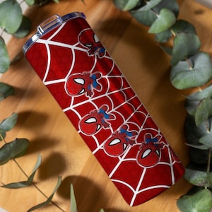 Może przedstawiać: Czerwono-biały kubek z motywem Spider-Mana. Kubek ma czerwone tło z białym wzorem pajęczyny i kilkoma grafikami twarzy Spider-Mana. Cylindryczny kubek ma srebrną pokrywę.