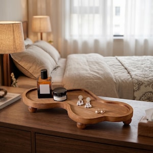 May include: An irregular, cloud-shaped wooden tray with a smooth, natural finish, supported by three small, rounded ball feet. The tray holds a rectangular amber glass bottle with a black cap, a small black jar, two dangling pearl earrings, and two pearl studs.