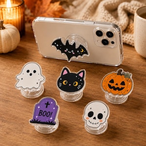 Op de afbeelding: Een verzameling van zes ronde telefoonhouders met Halloween-thema, waaronder een zwarte vleermuis, een wit spookje, een zwart kattengezicht, een oranje pompoen, een paarse grafsteen met de tekst BOO! en een witte schedel met roze wangen.