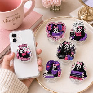 May include: A collection of six acrylic phone grips featuring a black ghost character wearing a pink bow and reading books. Each grip displays unique designs with text including Books are my Therapy, Smut Reader Club, Just One More Chapter, and Spicy Stories.