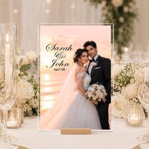 May include: A rectangular acrylic wedding sign featuring a photograph of a couple at sunset. The text reads Sarah & John August 7, 2026. The sign is supported by a light-colored wooden base and displays a bride in a white gown and a groom in a black suit.