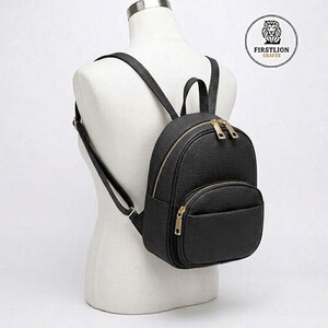 Puede incluir: Una mochila negra de cuero sintético con cremalleras y herrajes dorados. La mochila pequeña tiene forma redondeada con un bolsillo frontal y correas ajustables. La mochila se exhibe en un maniquí blanco.