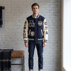 Op de afbeelding: Een marineblauw varsity-jack met crèmekleurige mouwen, voorzien van diverse geborduurde patches op de borst en mouwen, een drukknoopsluiting en gestreepte ribgebreide boorden bij de kraag, mouwen en zoom, gecombineerd met een donkerblauwe spijkerbroek.