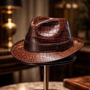 May include: A brown fedora hat featuring a textured crocodile skin pattern across the crown and brim. The hat includes a smooth, dark brown leather band encircling the base of the crown, finished with a small, rectangular buckle detail on the side.