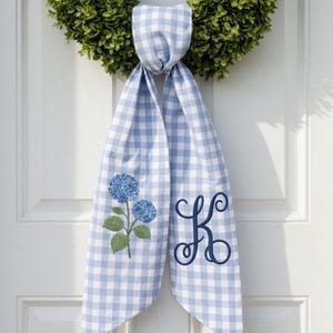 Puede incluir: Una corona verde cuelga de una puerta blanca, con una cinta de cuadros azul y blanco. La cinta presenta un bordado de hortensia azul y una letra cursiva "K".