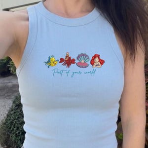 May include: A light blue ribbed tank top featuring a graphic print of a yellow fish, a red crab, a purple seashell with a pearl, and a red-haired character. Below the illustrations, the text Part of your world is printed in a cursive script.