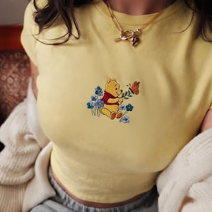 May include: A yellow t-shirt with an embroidered design of Winnie the Pooh holding a sprig with a butterfly, surrounded by blue flowers. The shirt is short-sleeved.