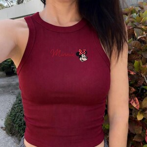 May include: A burgundy ribbed vest top with the name "Minnie" in cursive and a Minnie Mouse graphic. The sleeveless top has a round neckline and is made of a soft, stretchy material. The Minnie Mouse graphic includes a red bow with white polka dots.