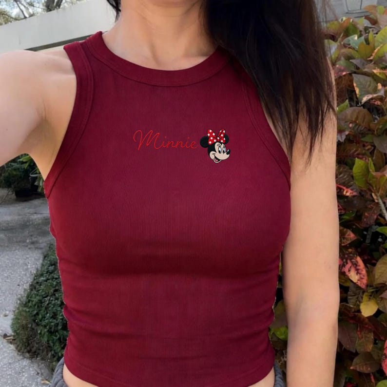 May include: A burgundy ribbed vest top with the name "Minnie" in cursive and a Minnie Mouse graphic. The sleeveless top has a round neckline and is made of a soft, stretchy material. The Minnie Mouse graphic includes a red bow with white polka dots.