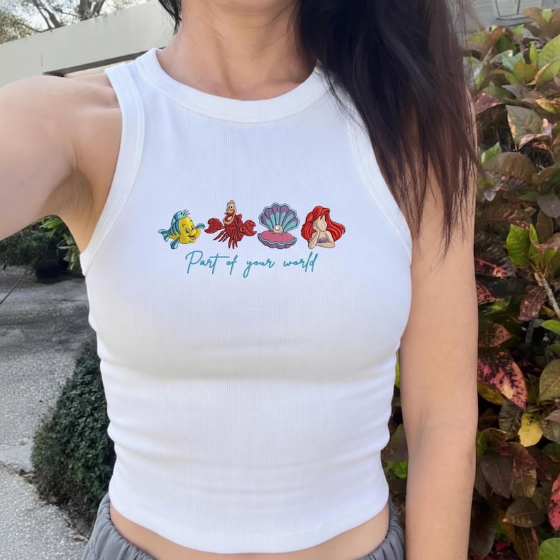 May include: A white ribbed tank top featuring a graphic print of a yellow fish, a red crab, a purple seashell with a pearl, and a red-haired character. Below the characters, the text Part of your world is printed in a teal cursive script font.
