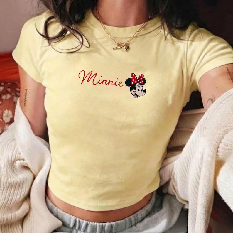 May include: A pale yellow t-shirt with the name "Minnie" in red script and a graphic of Minnie Mouse with a red and white polka dot bow. The top has short sleeves and a crew neck.