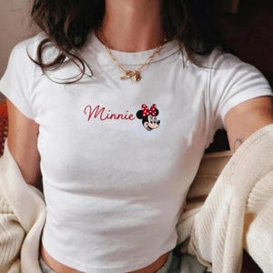 May include: White t-shirt featuring the word "Minnie" in red script, alongside a cartoon image of Minnie Mouse with a red polka dot bow. The top has short sleeves and a crew neck.