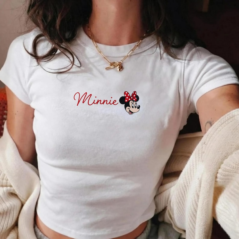 May include: White t-shirt featuring the word "Minnie" in red script, alongside a cartoon image of Minnie Mouse with a red polka dot bow. The top has short sleeves and a crew neck.