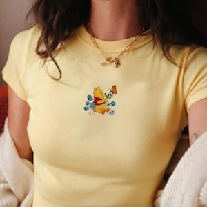 May include: Pale yellow t-shirt with a Winnie the Pooh design. The graphic depicts Pooh Bear with flowers and a butterfly. The top has short sleeves and a round neck. A gold necklace is worn.