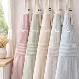 May include: Five striped aprons in pastel colors hang on a wooden rack. Each apron has a different color, including pink, green, beige, light blue, and a light blue with a small white tag. The aprons have a pocket and adjustable straps.