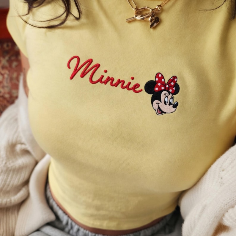 May include: Pale yellow t-shirt featuring the embroidered word "Minnie" in red script, alongside an embroidered Minnie Mouse graphic with a red polka dot bow. A gold necklace is visible.