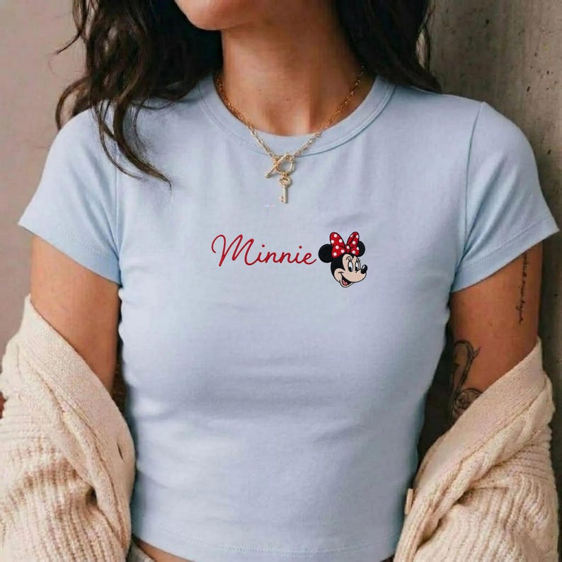 May include: Pale blue t-shirt with the word "Minnie" in red script and a Minnie Mouse graphic with a red polka dot bow. The top has short sleeves and a crew neck.