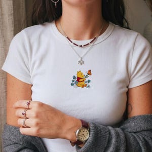 May include: White t-shirt with an embroidered design of Winnie the Pooh, a butterfly, and blue flowers. The cartoon character is holding a flower. The design is centered on the shirt.