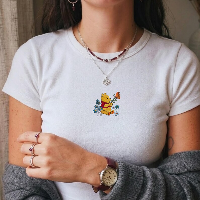 May include: White t-shirt with an embroidered design of Winnie the Pooh, a butterfly, and blue flowers. The cartoon character is holding a flower. The design is centered on the shirt.