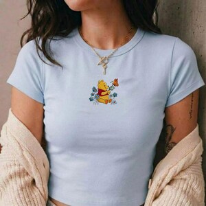 May include: Pale blue crop top with a Winnie the Pooh design. The cartoon bear is holding a flower and observing a butterfly. The top has short sleeves and a round neck.
