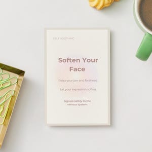 May include: A card with the text "Soften Your Face" is centered on a white surface. The card is surrounded by a gold tray with green paper clips, a green mug of coffee, and a cookie.