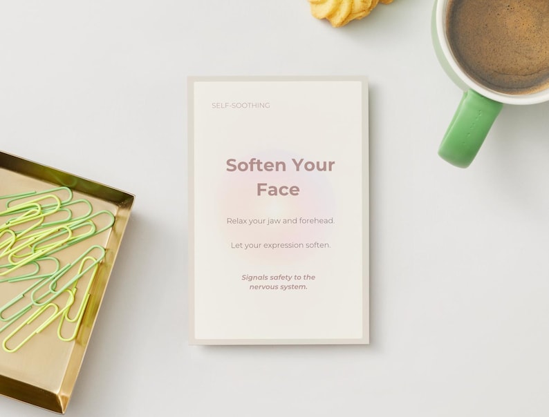 May include: A card with the text "Soften Your Face" is centered on a white surface. The card is surrounded by a gold tray with green paper clips, a green mug of coffee, and a cookie.
