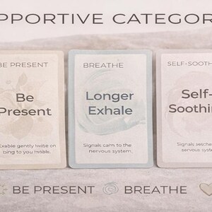 May include: Six supportive category cards with text including "Grounding", "Be Present", "Breathe", "Self-Soothing", and "Movement". Each card has a unique design and message. A lit candle sits to the right of the cards.