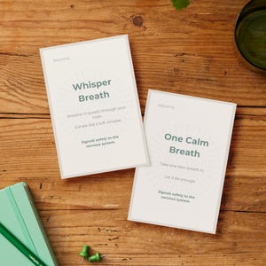May include: Two white cards with the word "Breathe" at the top, featuring the phrases "Whisper Breath" and "One Calm Breath." A green notebook, pencil, and push pins are on a wooden surface.