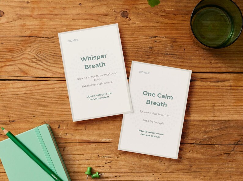 May include: Two white cards with the word "Breathe" at the top, featuring the phrases "Whisper Breath" and "One Calm Breath." A green notebook, pencil, and push pins are on a wooden surface.