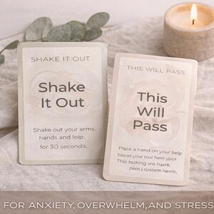 May include: Two rectangular cards with text, "Shake It Out" and "This Will Pass", are displayed. The "Shake It Out" card instructs to shake arms and hands for 30 seconds. The "This Will Pass" card has instructions. A lit candle and greenery are in the background. For anxiety, overwhelm, and stress.