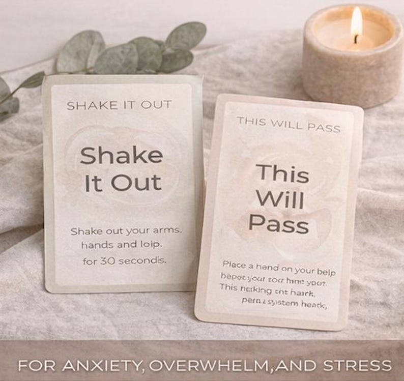 May include: Two rectangular cards with text, "Shake It Out" and "This Will Pass", are displayed. The "Shake It Out" card instructs to shake arms and hands for 30 seconds. The "This Will Pass" card has instructions. A lit candle and greenery are in the background. For anxiety, overwhelm, and stress.