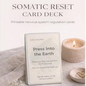 May include: A card deck titled "SOMATIC RESET CARD DECK" with the text "Printable nervous system regulation cards." One card reads "Press Into the Earth." A lit candle and small dish with paper clips are also visible.