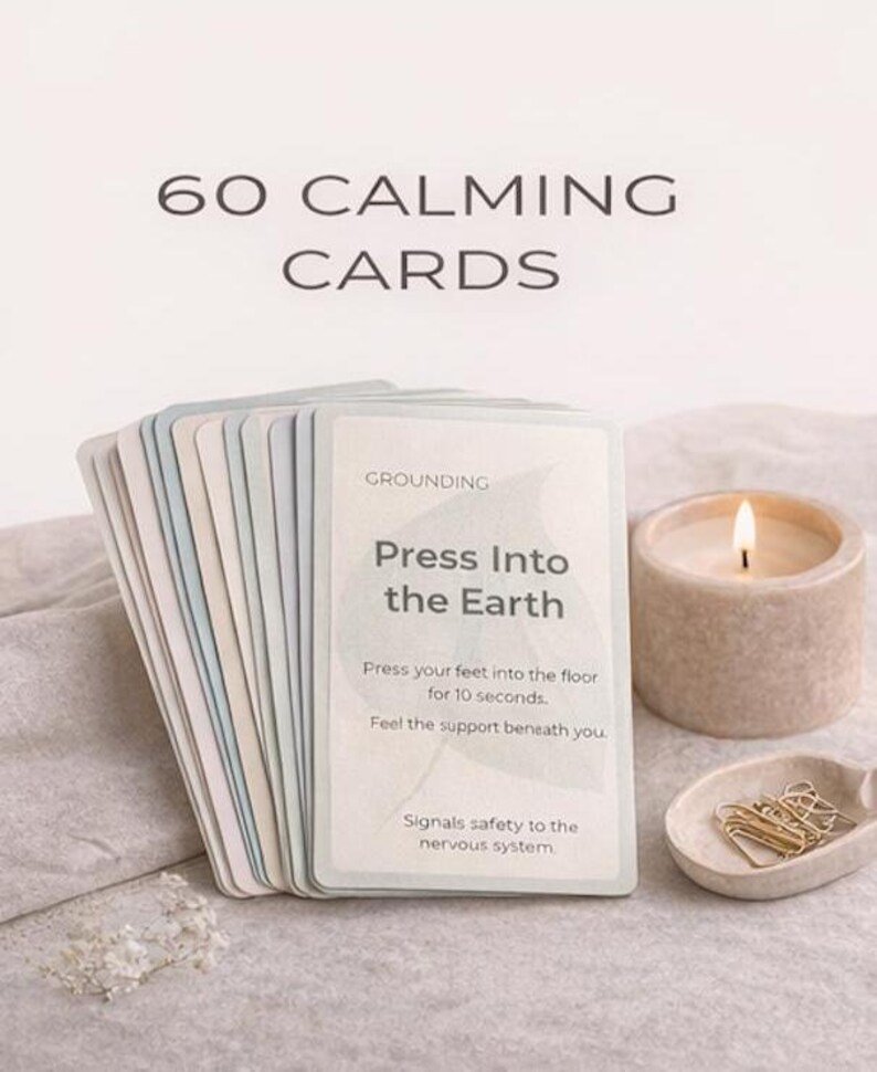 May include: A stack of 60 calming cards with the text "Press Into the Earth" and instructions. A lit candle and a small dish with paper clips are next to the cards. The text "60 CALMING CARDS" is at the top.