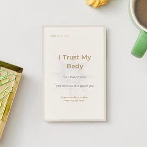 May include: A card with the text "I Trust My Body" and supporting text, next to a gold tray of green and yellow paperclips, a green-handled mug of coffee, and a cookie on a white surface.