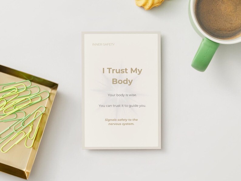 May include: A card with the text "I Trust My Body" and supporting text, next to a gold tray of green and yellow paperclips, a green-handled mug of coffee, and a cookie on a white surface.
