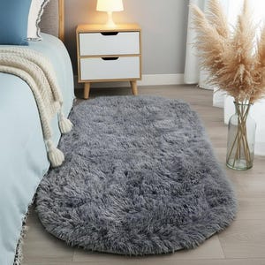 May include: A gray, oval-shaped, fluffy rug. The rug is made of soft, shaggy material. The rug is on a hardwood floor. A bed with a blue comforter and a white nightstand are in the background.